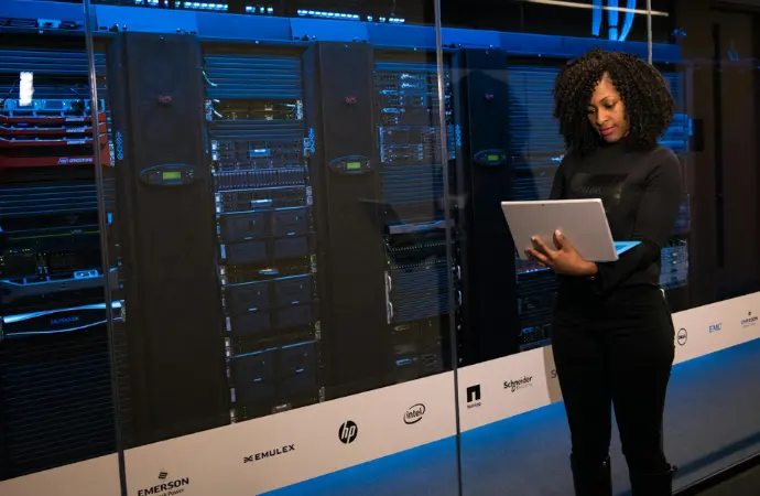 A woman stands confidently in front of a server rack, representing ServiceNow SecOps and cybersecurity solutions.